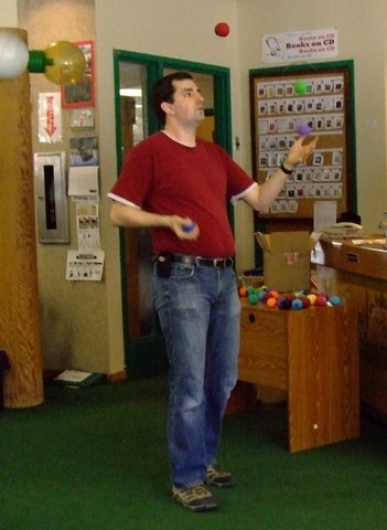 A tall man in a red shirt and jeans juggling three balls